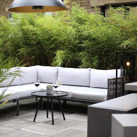 Middle Eight - Covent Garden - Preferred Hotels And Resorts London Bagian luar foto The photo shows an outdoor seating area featuring a modern gray sectional sofa with plush cushions. In front of the sofa is a small, round black table with two glasses of wine on it, accompanied by a decorative plant or vase. Surrounding the area are
